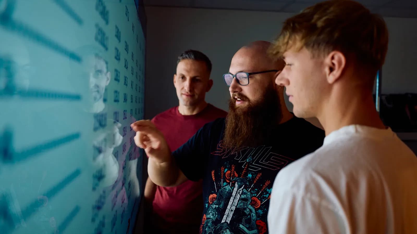 Drei Mitarbeiter besprechen sich vor einer großen Monitoring-Wand mit Systemdaten.