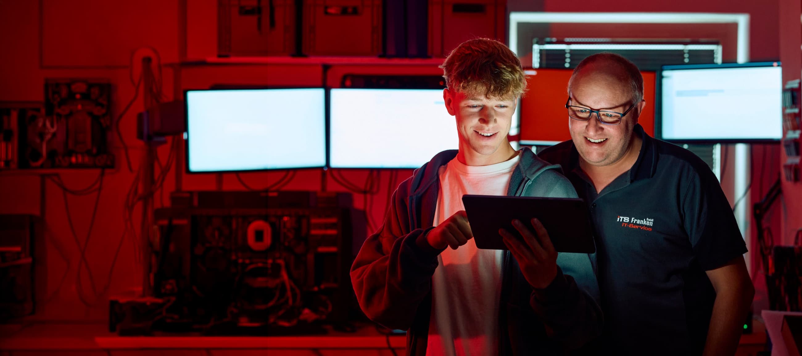 Zwei ITS-Mitarbeiter besprechen gemeinsam Systemdaten auf einem Tablet in einem rot beleuchteten Serverraum.