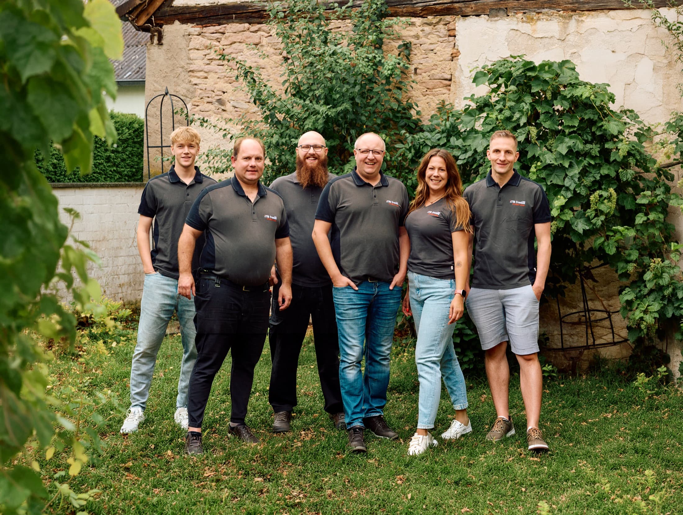 Das Team der ITS Franken IT-Service GmbH steht gemeinsam im Freien vor einer Natursteinmauer.